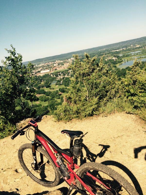 A mountain bike rests on a dirt trail overlooking a lush green valley and a small town in the distance, under a clear blue sky. Hixon Forest mountain bike trail.