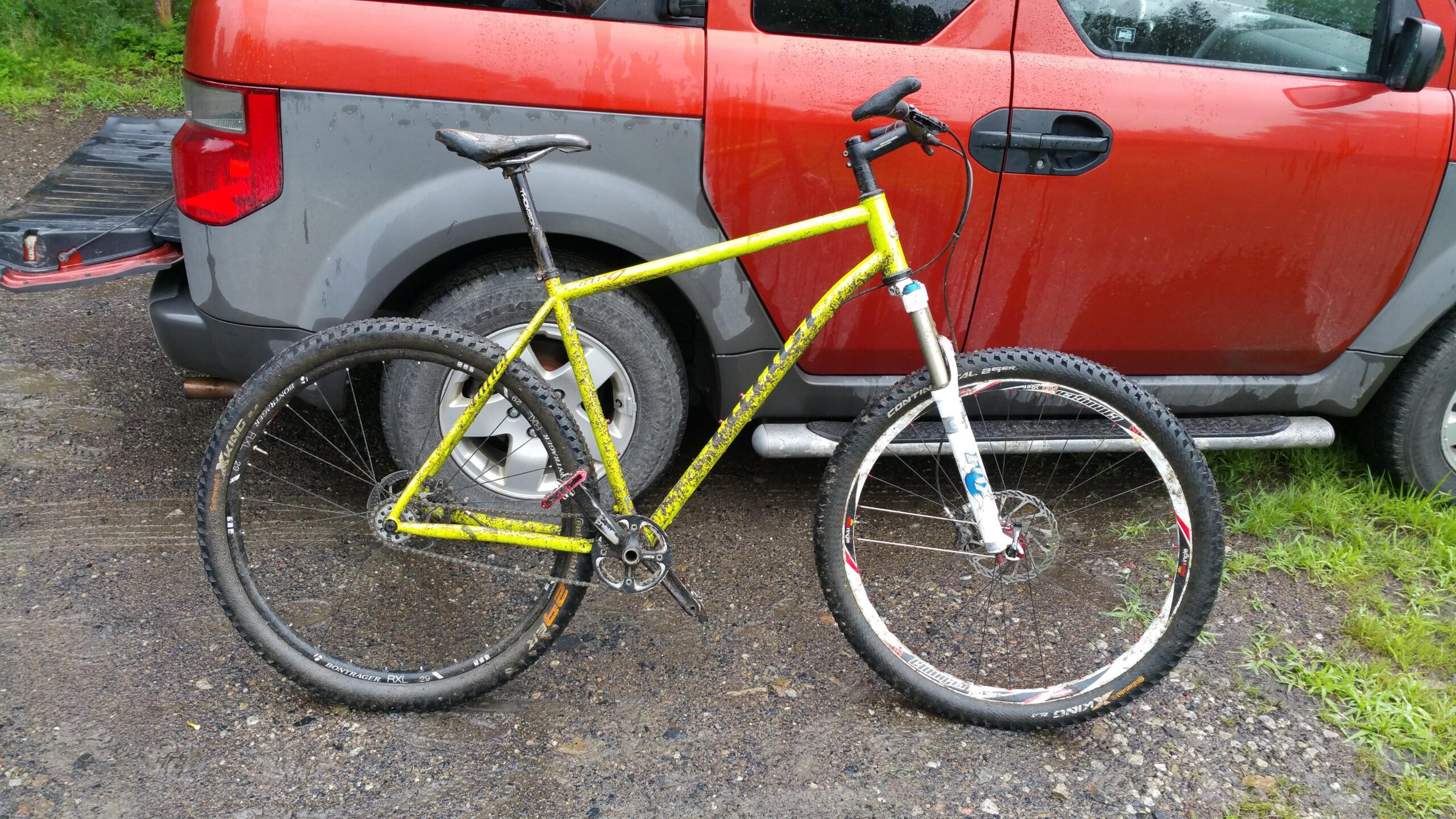 Niner S.I.R. 9: A bright yellow mountain bike is parked next to a red vehicle with a gray rear section. The bike has a white front fork and is slightly muddy, indicating recent use on rough terrain. The background features a gravel surface and some green grass.
