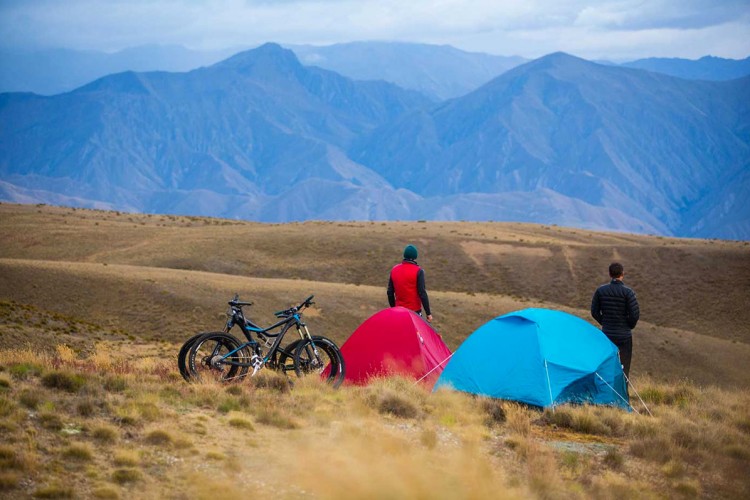 Two people stand near colorful tents on a grassy hilltop, overlooking a vast mountain range in the background. Two mountain bikes rest beside the red and blue tents, with a dramatic sky filled with clouds above. The scene captures a sense of adventure and the beauty of the outdoors.