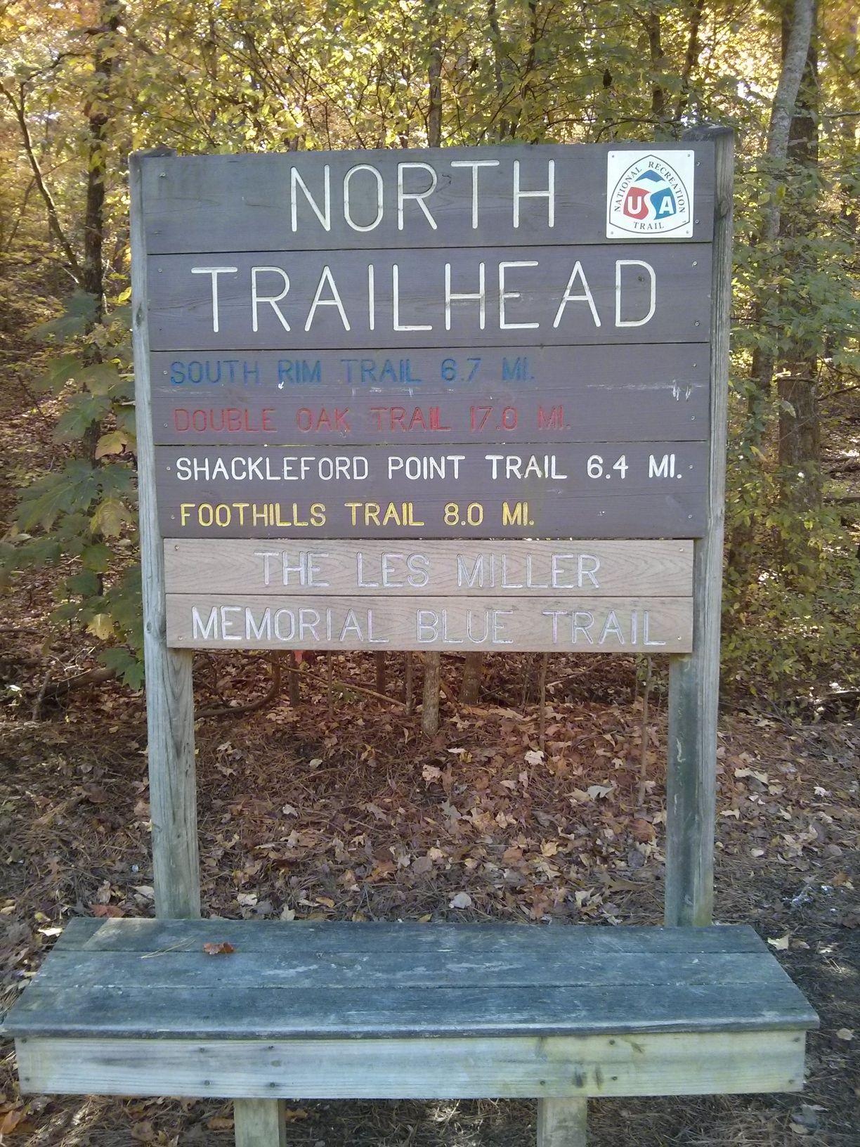 Signpost at a trailhead displaying various hiking trails and their distances, including South Rim Trail (6.7 miles), Double Oak Trail (17.0 miles), Shackleford Point Trail (6.4 miles), and Foothills Trail (8.0 miles). The sign also mentions The Les Miller Memorial Blue Trail. The surrounding area features trees and fallen leaves. Oak Mountain State Park mountain bike trail.