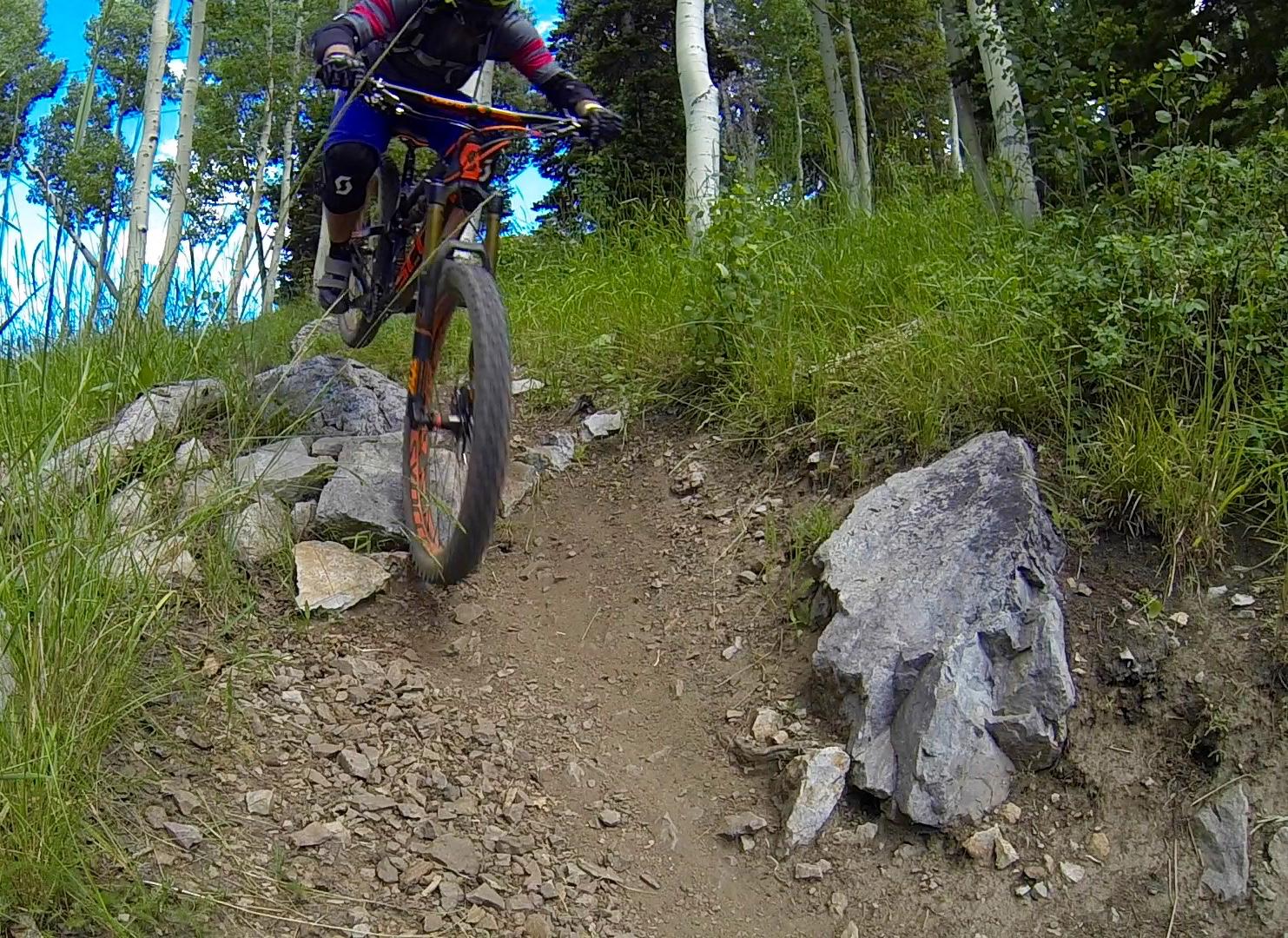 A mountain biker navigating a rocky trail, airborne over a large rock, with green grass and trees in the background under a blue sky. Deer Valley Resort Bike Park mountain bike trail.