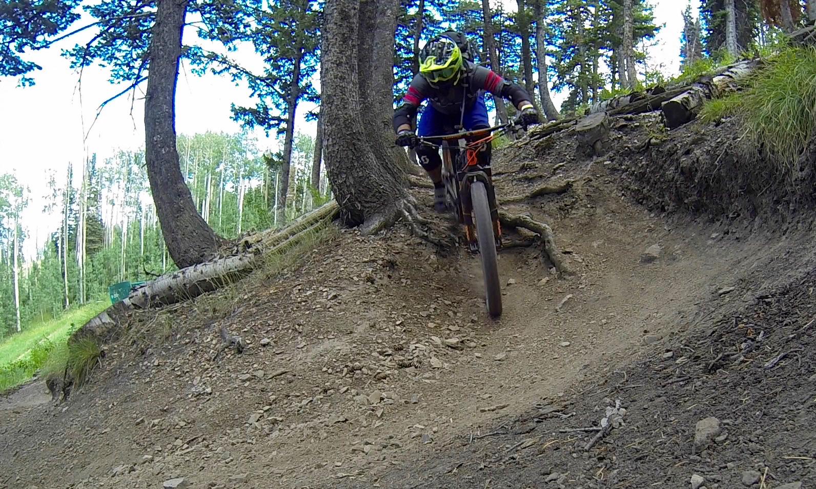 A mountain biker navigating a dirt trail surrounded by trees, leaning forward as they approach a turn. The terrain shows some elevation changes and roots, highlighting the challenging nature of the trail. Deer Valley Resort Bike Park mountain bike trail.
