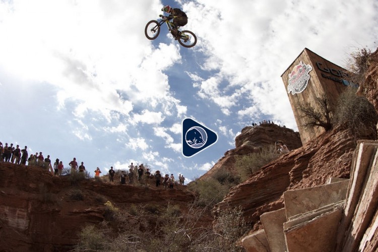 A mountain biker performs a mid-air jump over a rocky canyon during an event, with a crowd of spectators watching from the edge of the cliff above. The sky is partly cloudy, and a large wooden structure with branding is visible in the background.