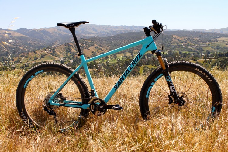 A bright blue mountain bike parked in tall grass, with rolling hills and trees in the background under a clear sky. The bike features a lightweight frame and rugged tires, suggesting it is designed for outdoor trails.