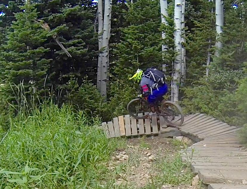 A mountain biker navigates a wooden trail through a lush forest, jumping off a small ramp. The biker wears a bright yellow helmet and a backpack, surrounded by tall green grass and trees. Deer Valley Resort Bike Park mountain bike trail.