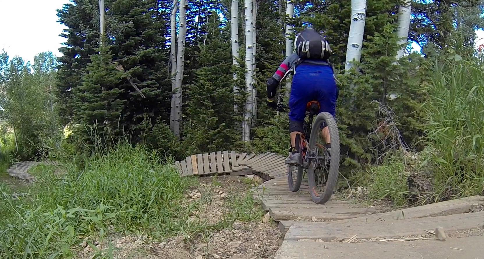 Mountain biker riding along a wooden trail surrounded by lush greenery and trees, capturing the movement through a natural landscape. Deer Valley Resort Bike Park mountain bike trail.