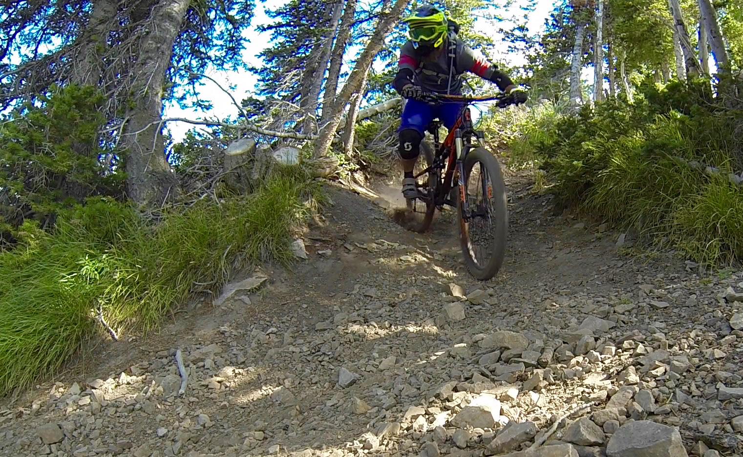 A mountain biker descending a rocky trail through a forested area, wearing protective gear and a bright-colored helmet. Sunlight filters through the trees, illuminating the rough terrain and vegetation along the path. Dust is kicked up from the rear wheel as the rider navigates the track. Deer Valley Resort Bike Park mountain bike trail.