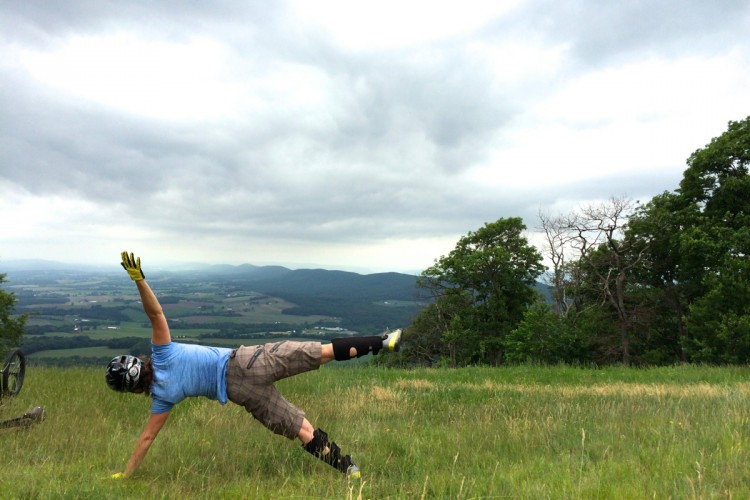A person in a blue shirt, shorts, and protective gear is performing a bike trick on a grassy hilltop with a scenic view of rolling hills and cloudy skies in the background. The individual is balancing on one hand while extending the other arm upward. A mountain bike is partially visible on the ground nearby.