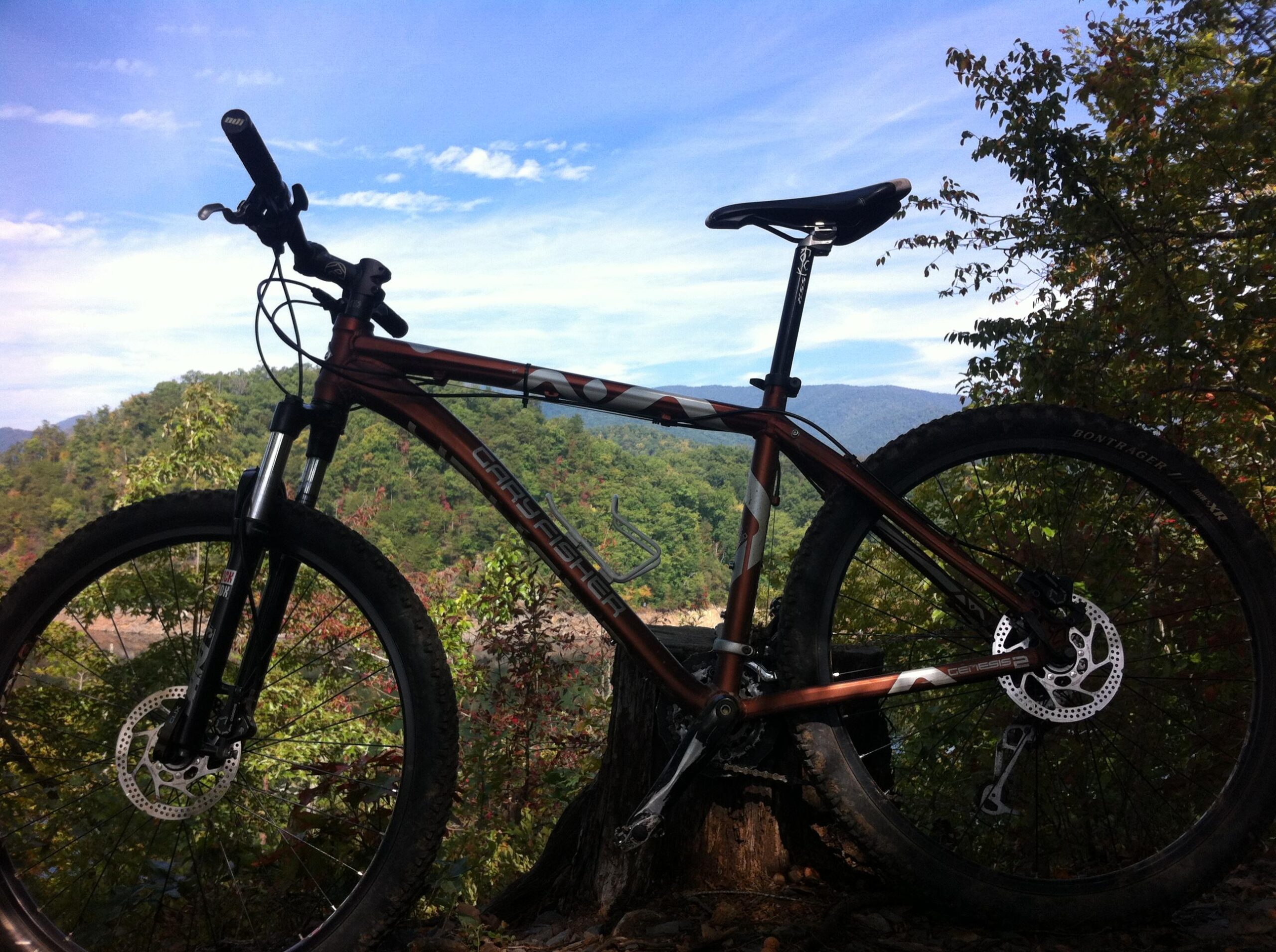Gary Fisher Piranha: Mountain bike resting on a tree stump with a scenic backdrop of green hills and a clear blue sky.