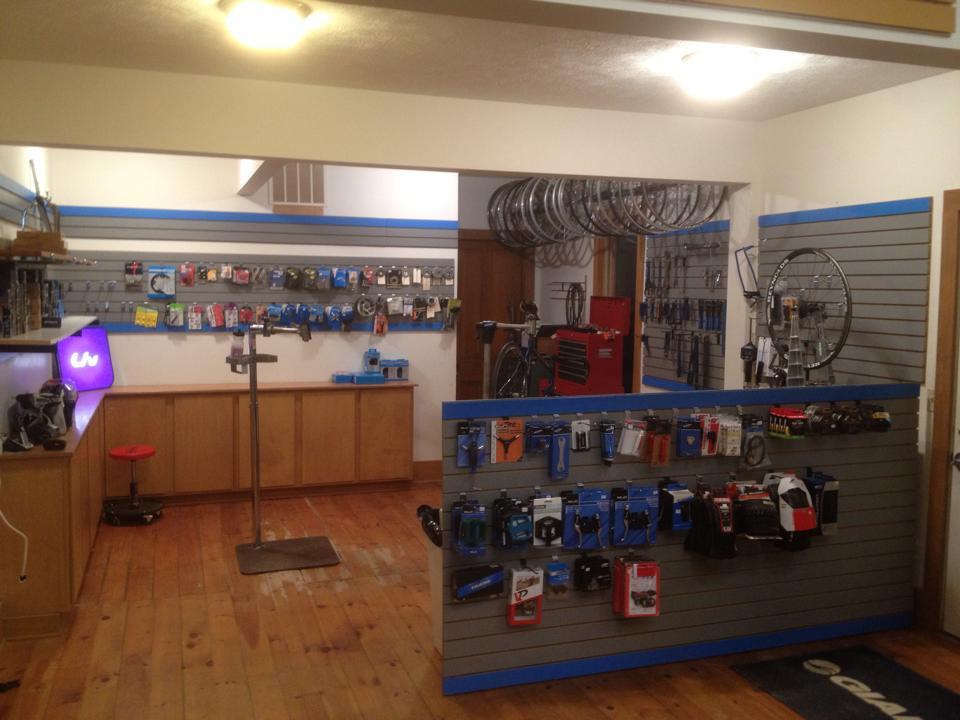 A bicycle repair shop interior featuring wooden flooring, a service area with a repair stand, and a wooden counter. The walls are lined with various bicycle parts and accessories, including tools and components displayed on pegboards. There are bike wheels hung on the walls, and a red toolbox is visible in the background. A small red stool is positioned near the service area.