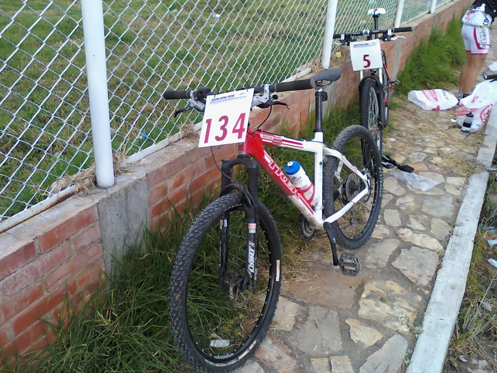 Trek Fuel EX 8: A close-up view of a mountain bike with race number 134 attached, leaning against a wall next to another bike displaying race number 5. The bikes are positioned near a grassy area and a chain-link fence, with a water bottle mounted on the frame of the first bike.