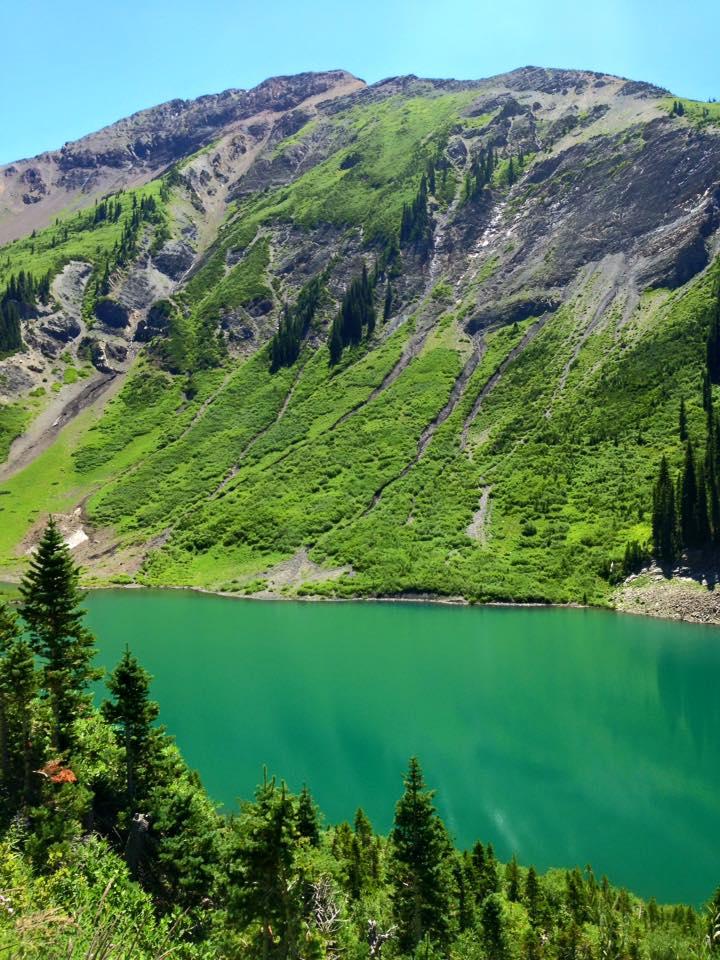 A vibrant mountain landscape featuring a turquoise lake surrounded by lush green vegetation and steep rocky slopes. Pine trees dot the foreground, leading up to the mountainous terrain under a clear blue sky. Trail 401 mountain bike trail.