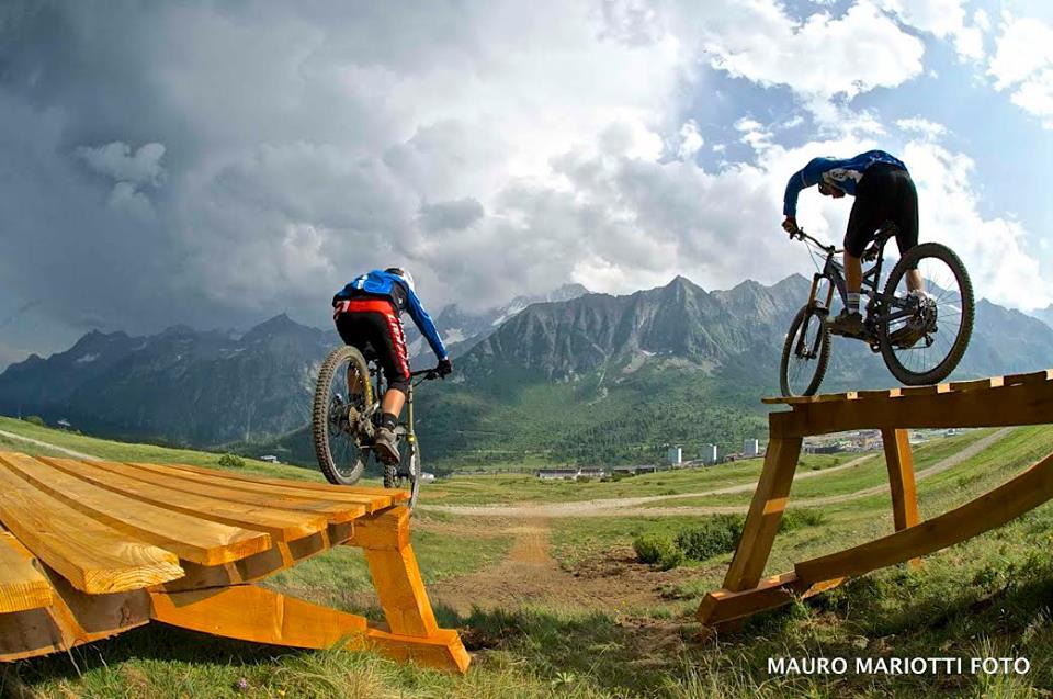 Two mountain bikers performing jumps on a wooden ramp with a mountainous landscape in the background, under a cloudy sky. Addable Bike Arena mountain bike trail.