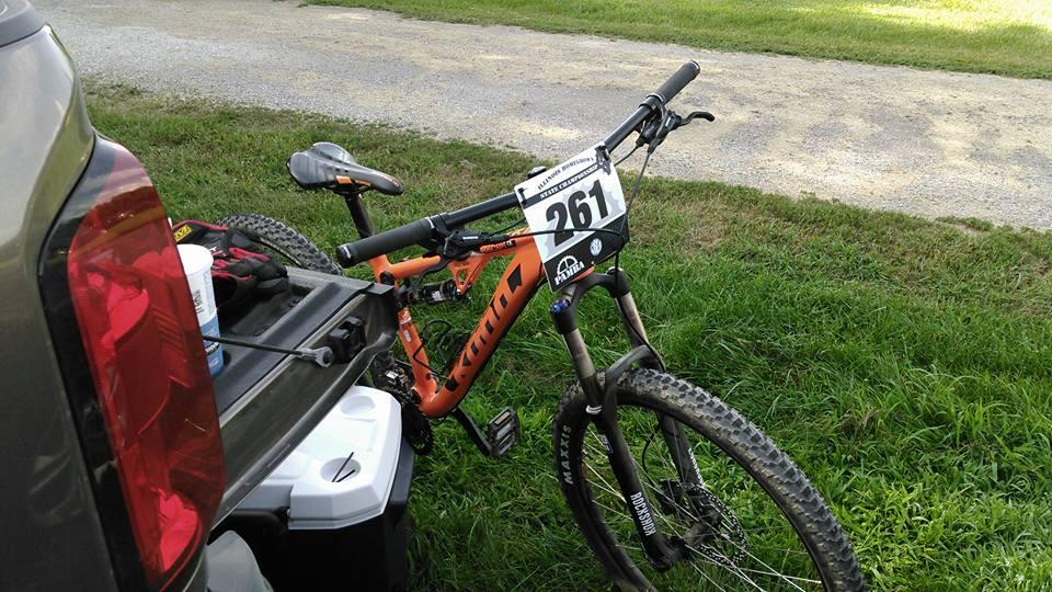 Kona Precept DL: A mountain bike with a race number 261 is resting against the tailgate of a truck, parked on grass near a gravel path. An assortment of biking gear, including a helmet and a water bottle, is visible in the truck bed.