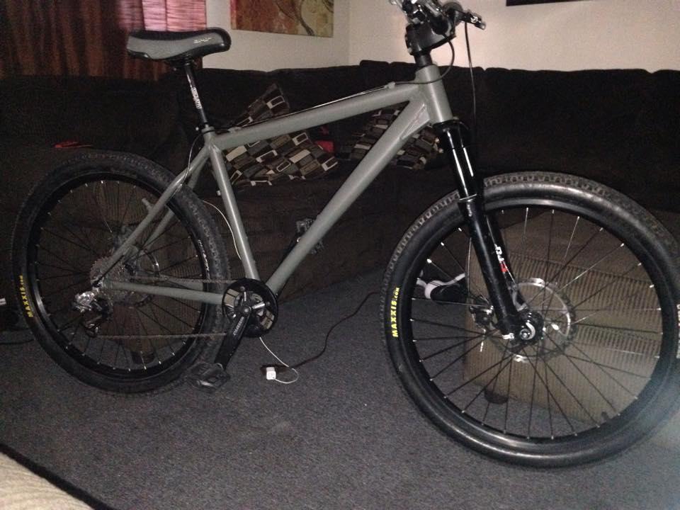Leader LD-516H: A grey mountain bike with black wheels and a sleek design, positioned on a carpeted floor in a living room. Visible in the background are a dark couch and a decorative wall piece.