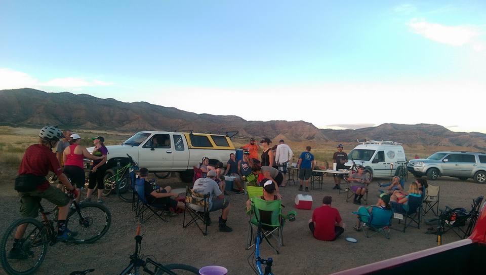 A group of people is gathered outdoors in a scenic area with mountains in the background. Some individuals are seated in camping chairs, while others are interacting or riding bicycles. Several vehicles are parked nearby, and the atmosphere appears lively and social, indicative of a community event or outdoor gathering.