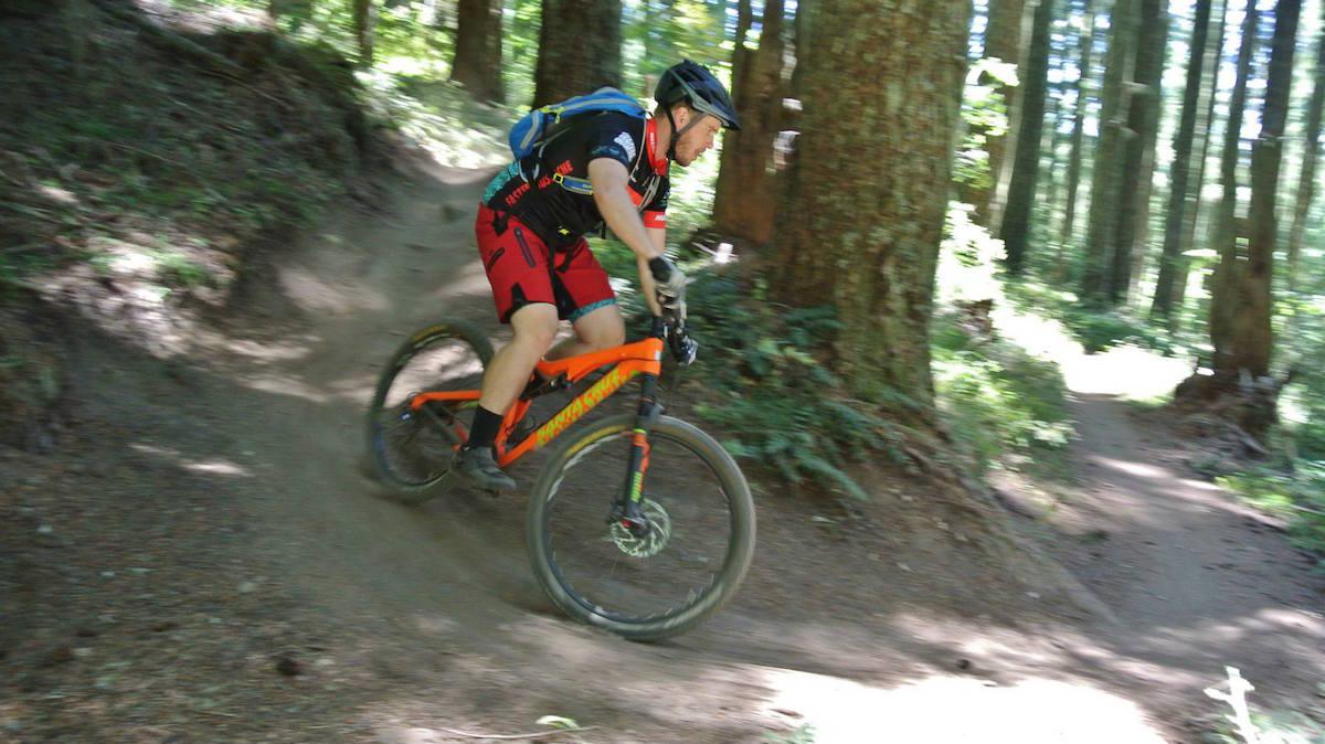 Santa Cruz 5010: A mountain biker rides along a winding dirt trail through a forest of tall trees, leaning into a turn on an orange bike with black accents. The scene captures the speed and agility of the rider, with dust kicked up behind them, surrounded by lush greenery.