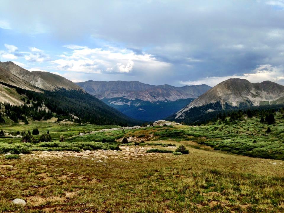 A scenic view of a mountainous landscape featuring rolling hills, scattered boulders, and lush green valleys under a cloudy sky. The peaks in the background are rugged and expansive, showcasing a mix of light and shadow. CDT: Alpine Tunnel mountain bike trail.