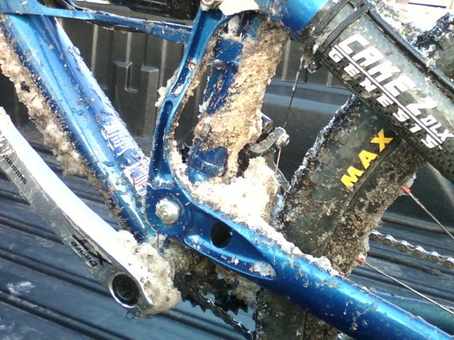 Close-up view of a muddy mountain bike frame showing thick clumps of dirt and grime around the bike's components, particularly near the bottom bracket and chainstay. The bike features a blue frame with black wheels and a partially obscured tire brand visible. The background is slightly blurred, suggesting it's in the bed of a truck. Big South Fork mountain bike trail.