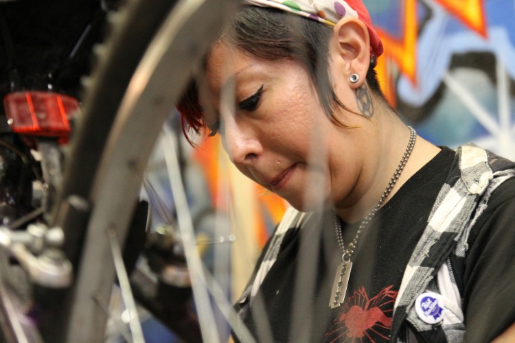 A close-up image of a person working on a bicycle, focused on their task. The individual has short hair with a colorful bandana, earrings, and visible tattoos. They are carefully handling a bike component, surrounded by blurred bicycle parts and a colorful graffiti background.