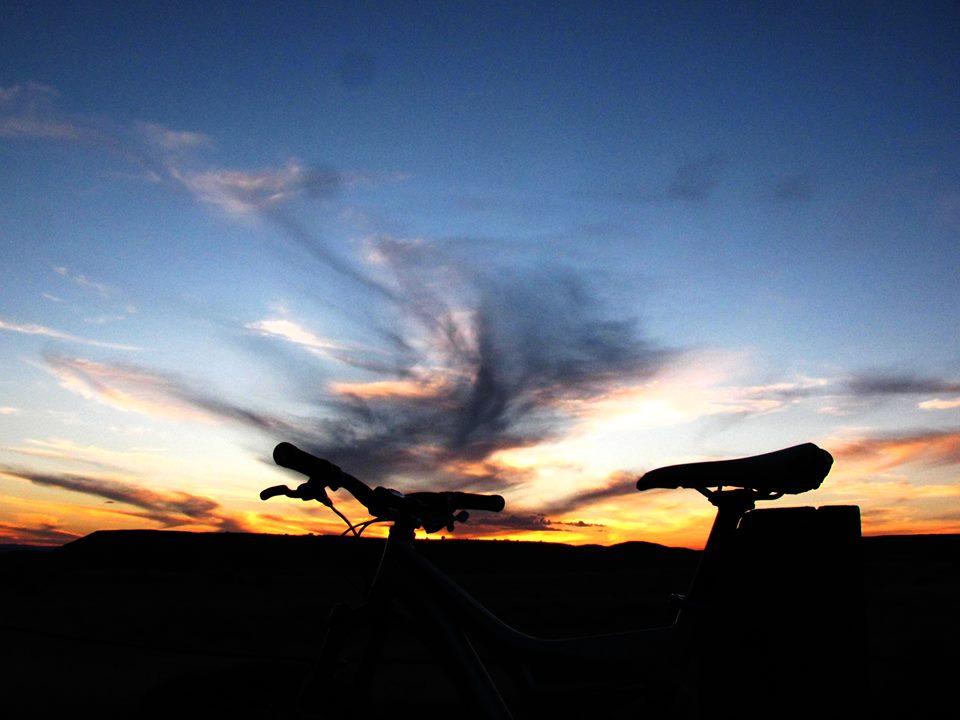 Silhouette of a bicycle against a colorful sunset sky with clouds. The horizon features vibrant hues of orange, pink, and blue.