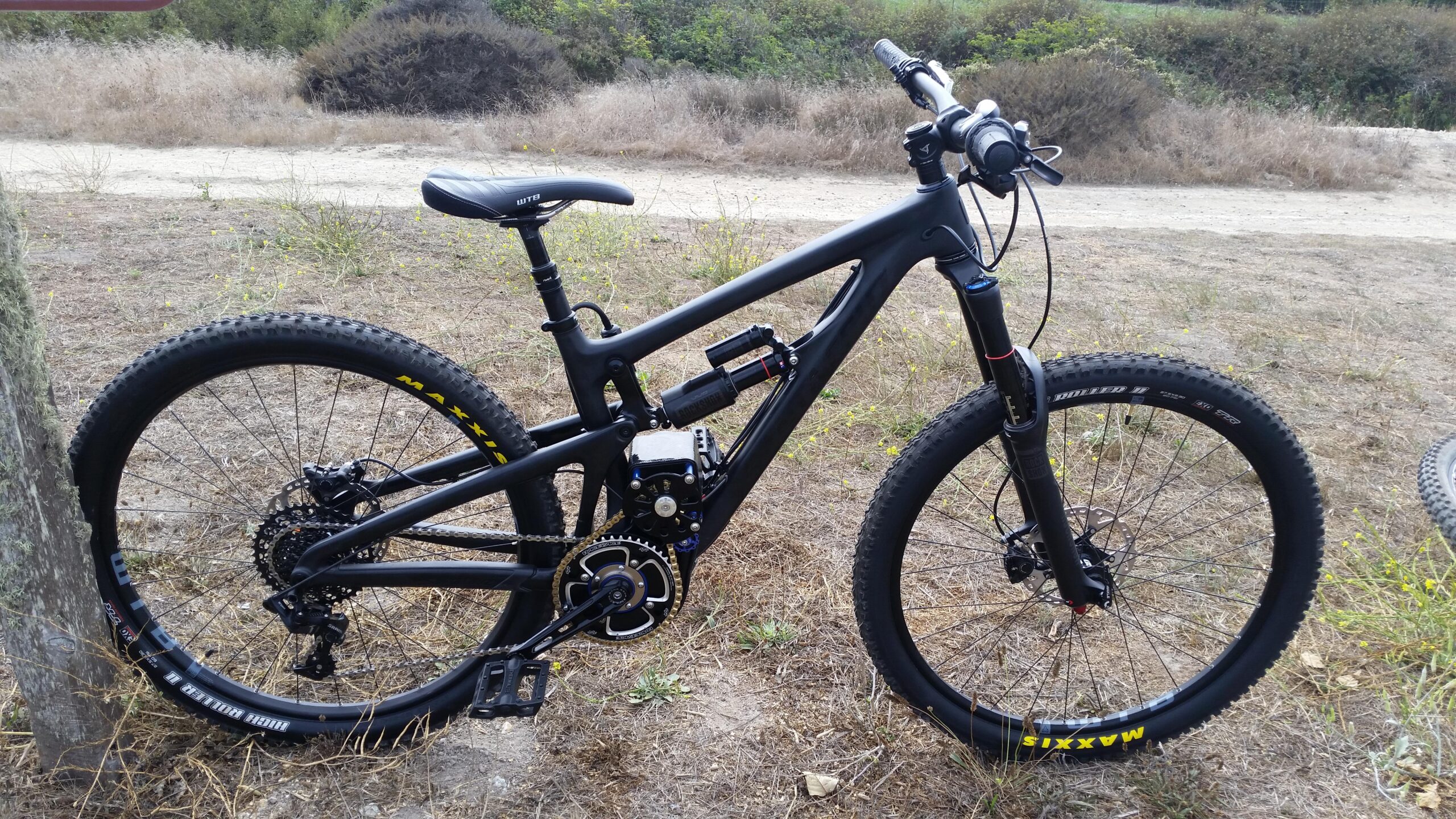 Santa Cruz Nomad: Black mountain bike leaning against a post, featuring knobby tires, a suspension system, and a chain drivetrain. The background consists of dry grass and shrubs, with a dirt path visible in the distance.