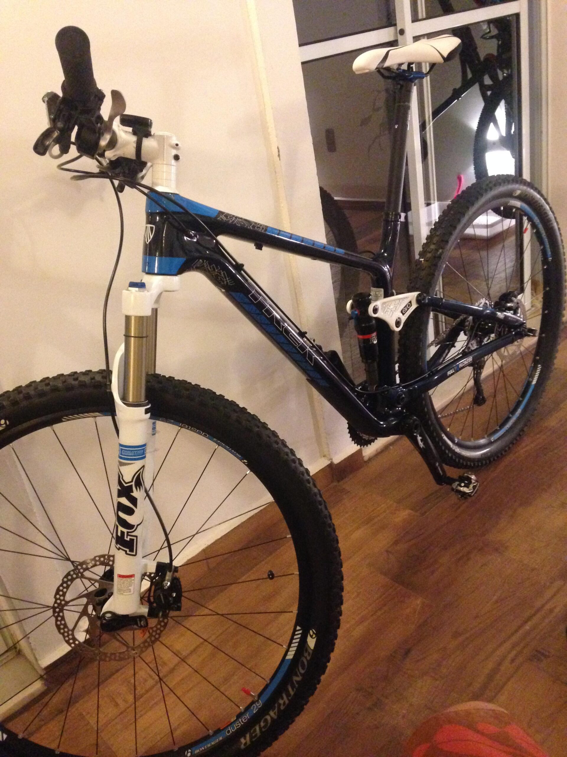 Trek Fuel EX 9.7 29: A mountain bike positioned against a wall, featuring a black and blue frame, front suspension fork, and disc brakes. The bike has thick, knobby tires suitable for off-road terrain, and the handlebars are equipped with gear shifters. Reflections from a nearby window and wooden flooring are visible in the background.