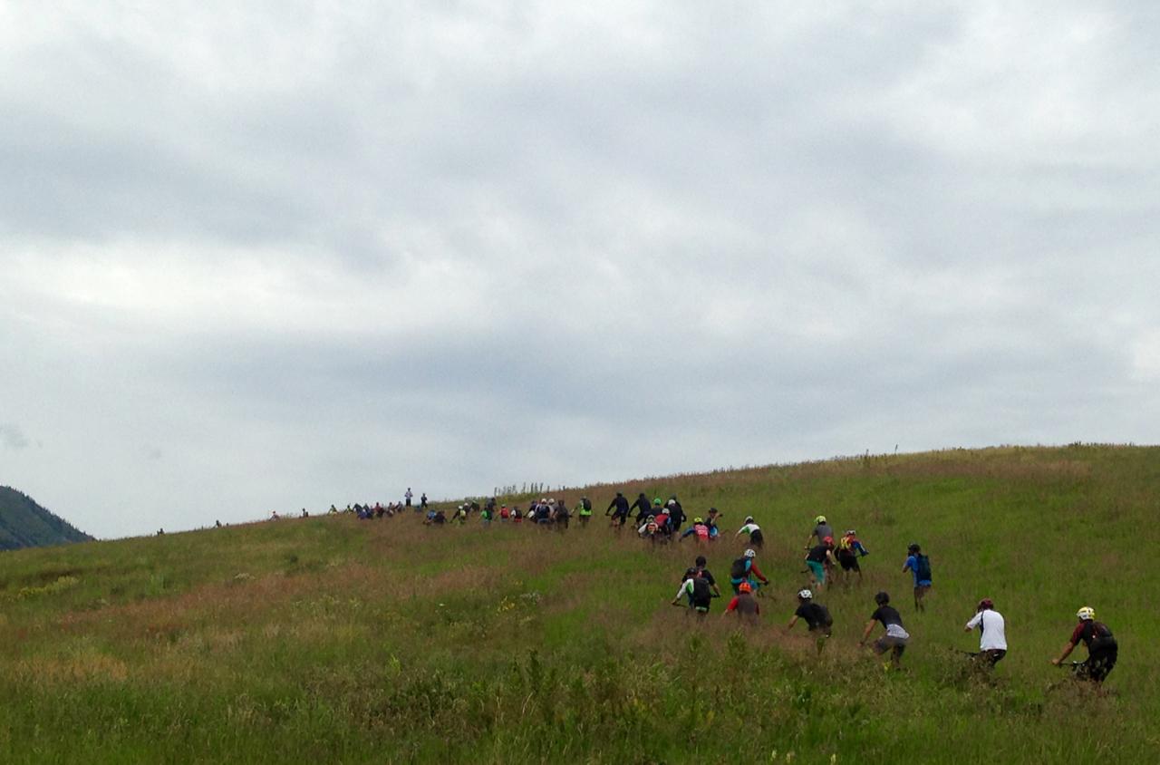 A group of cyclists ascending a grassy hill on a cloudy day, showcasing a mix of colorful biking attire against a backdrop of greenery. Lupine Trail mountain bike trail.