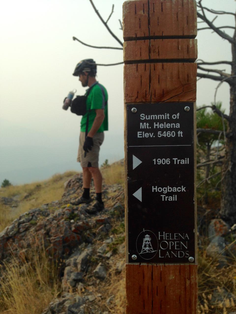 A cyclist in a green shirt stands on rocky terrain, near a wooden trail sign indicating the summit of Mt. Helena at an elevation of 5460 feet. The sign points to the 1906 Trail and Hogback Trail, with the background featuring a hazy landscape and dry grass. Mt Helena Ridge mountain bike trail.