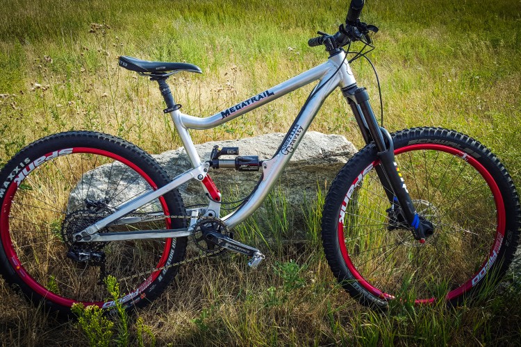 A mountain bike with a silver frame, red accents on the wheels, and thick tires, resting beside a rock in a grassy field.