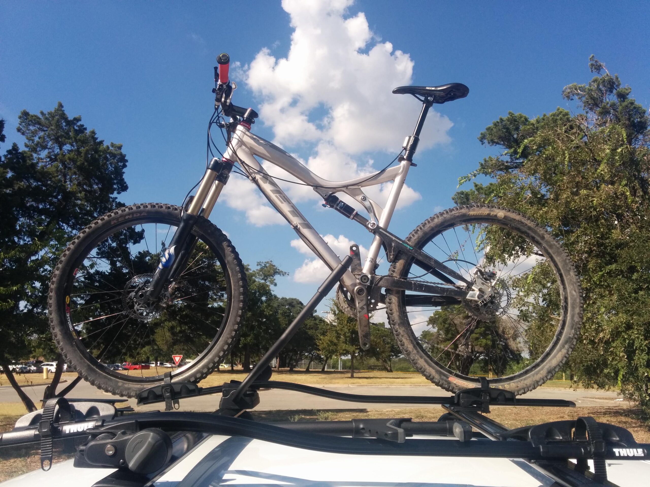 Yeti 575: A mountain bike mounted on the roof rack of a car, set against a blue sky with fluffy white clouds. The bike features knobby tires, suspension forks, and a bright red handlebar grip. Surrounding the scene are trees and an open area, suggesting a recreational outdoor environment.