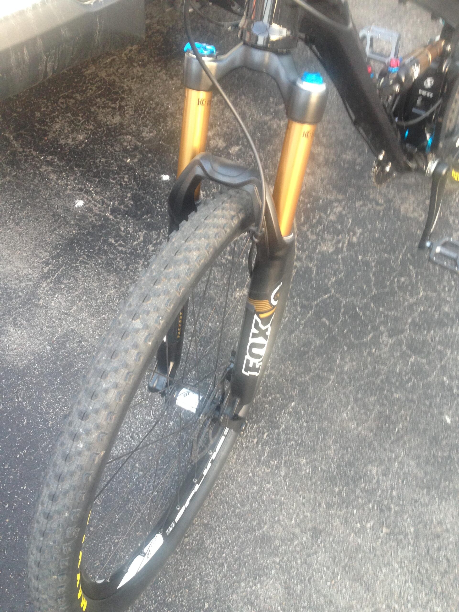 Yeti SB-75: Close-up view of a mountain bike's front suspension fork and wheel, featuring gold-colored suspension stanchions and a black tire on a textured asphalt surface. The bike is positioned at an angle, showcasing the fork and wheel details.