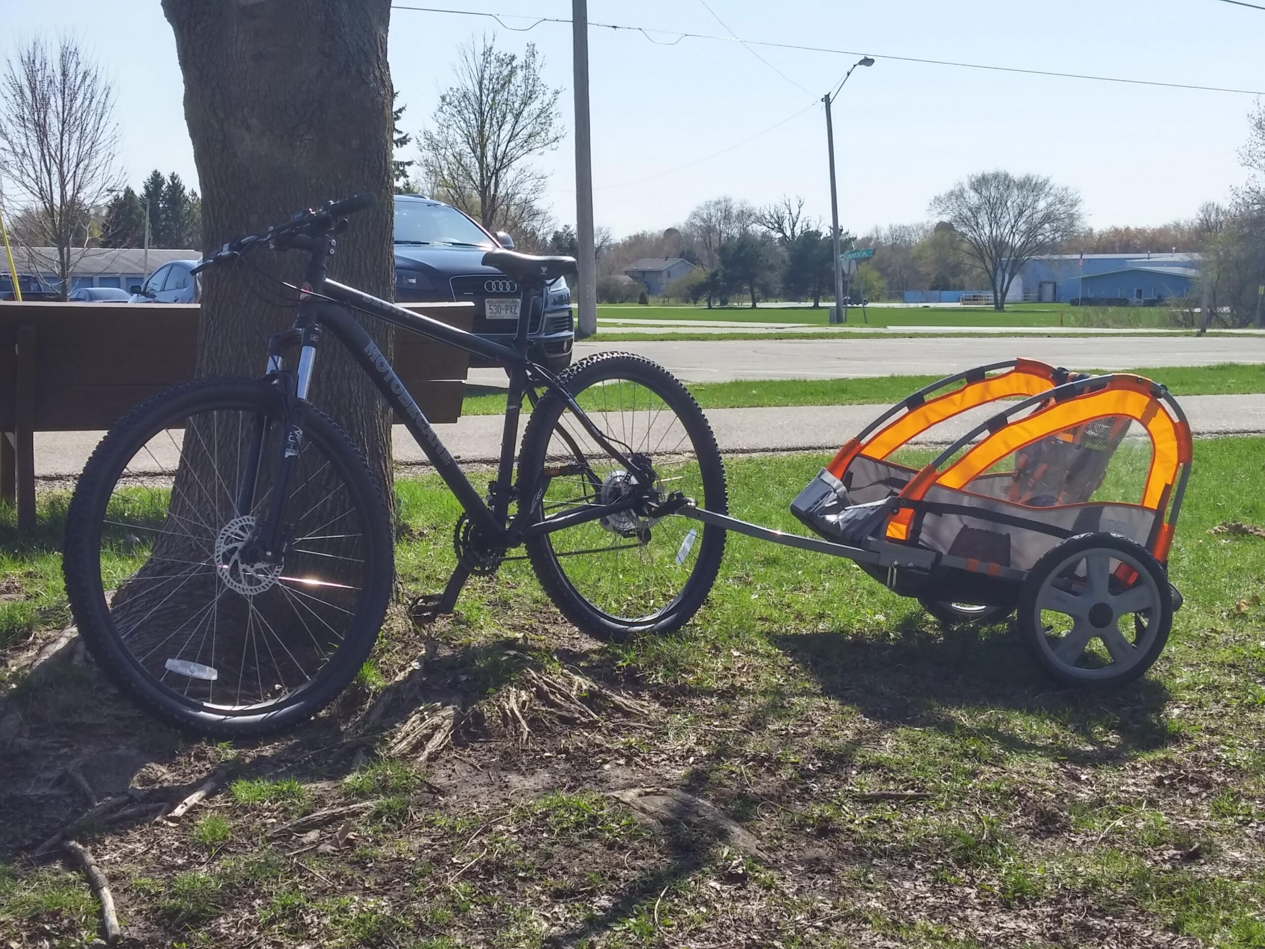 Motobecane Motobecane 529 HT: A black mountain bike parked next to a tree, connected to a colorful child bike trailer with orange accents. The scene is set in a grassy area with a clear sky, showcasing a suburban neighborhood in the background.