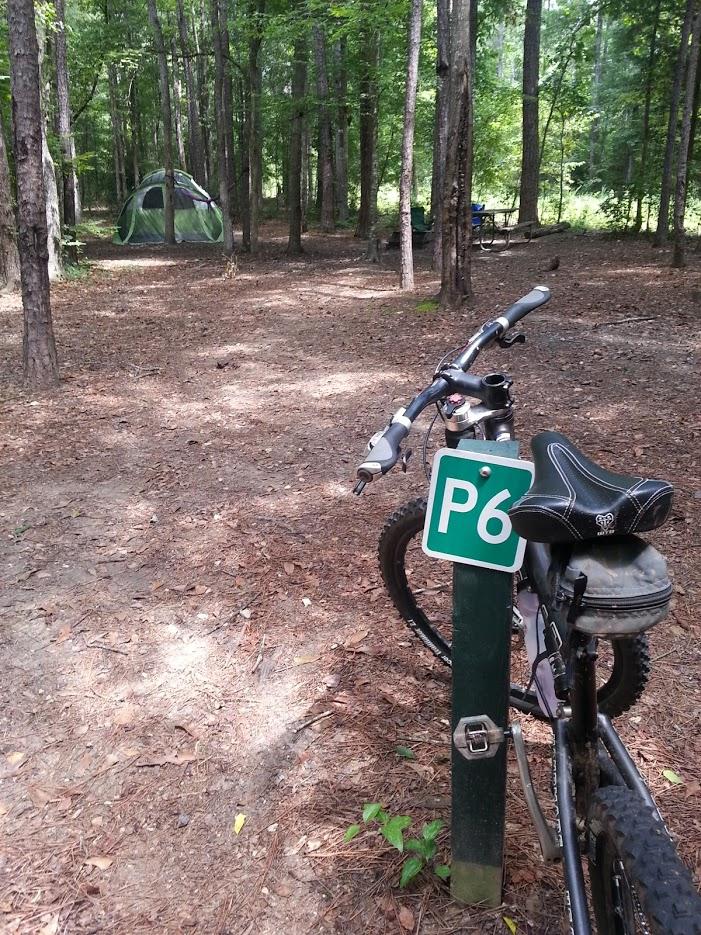 Bicycle parked next to a green sign marked "P6" in a wooded camping area, with a tent visible in the background among the trees. Pine needles and leaves cover the ground, creating a natural campsite atmosphere. Chewacla State Park mountain bike trail.