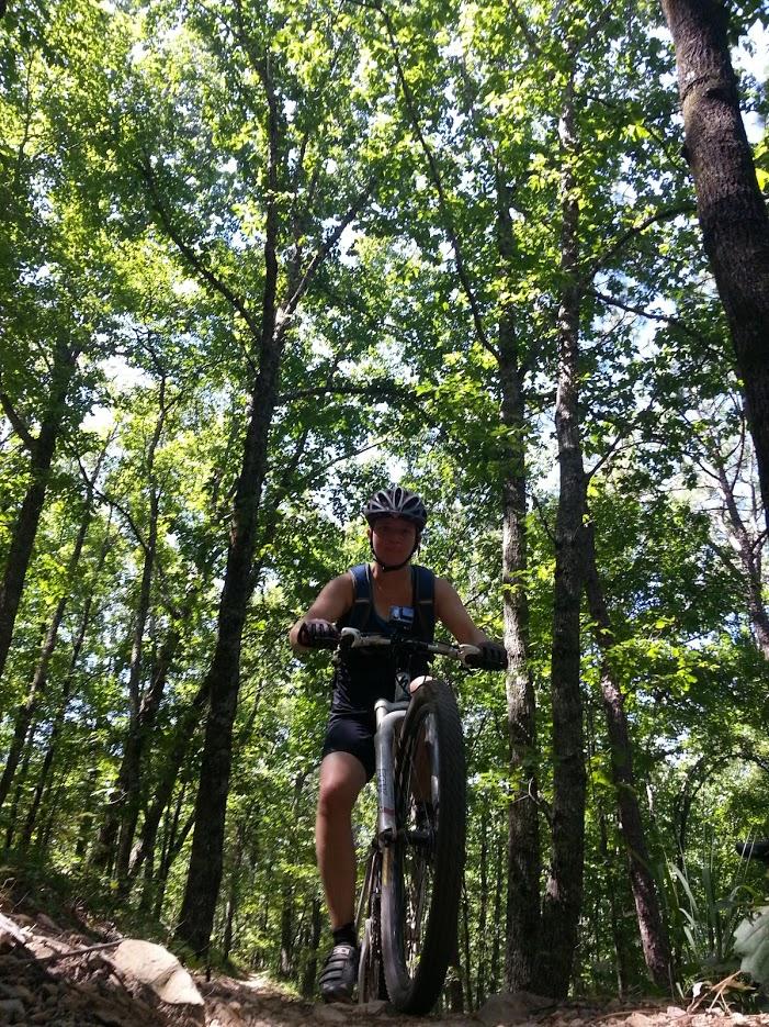 A person riding a mountain bike on a wooded trail, surrounded by tall trees and bright green foliage, wearing a helmet and athletic clothing. The sunlight filters through the leaves, creating a vibrant outdoor scene. Oak Mountain State Park mountain bike trail.