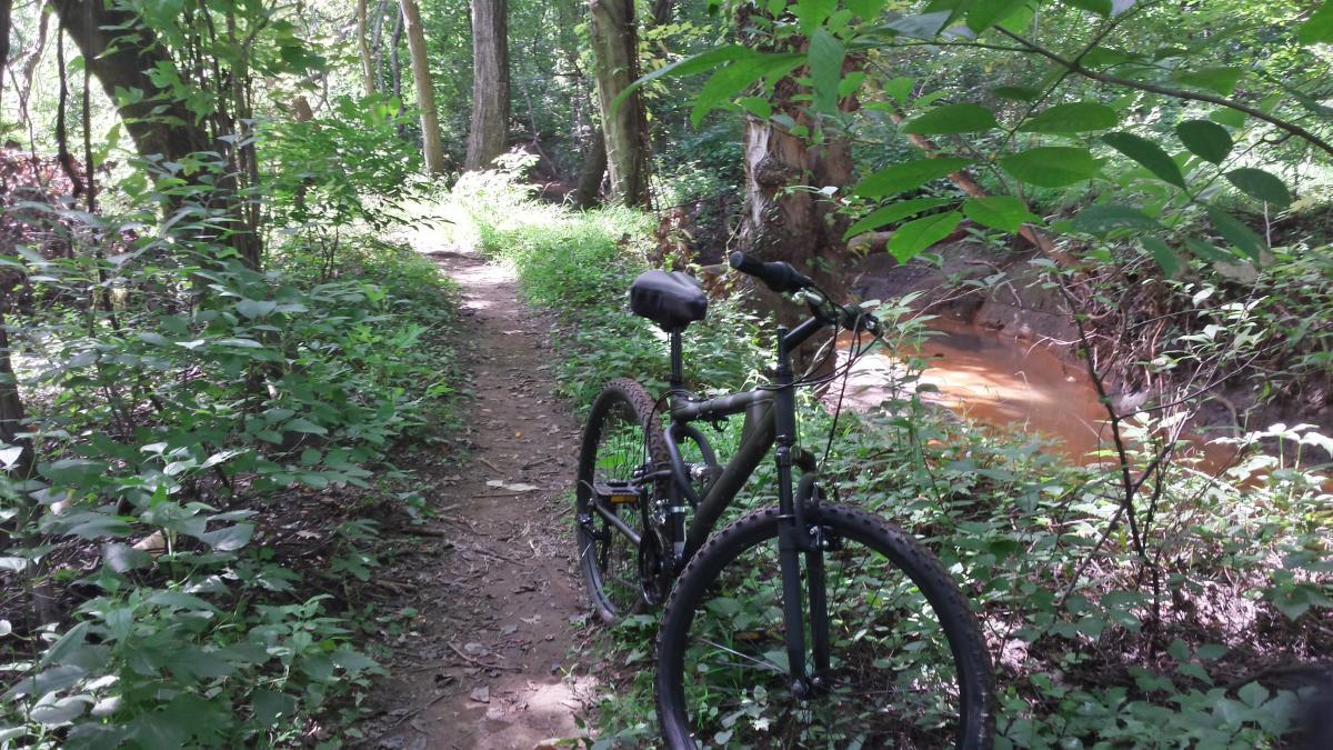 Hyper Havoc: A mountain bike parked beside a narrow dirt path in a lush green forest, surrounded by tall trees and dense underbrush, with a visible stream in the background.