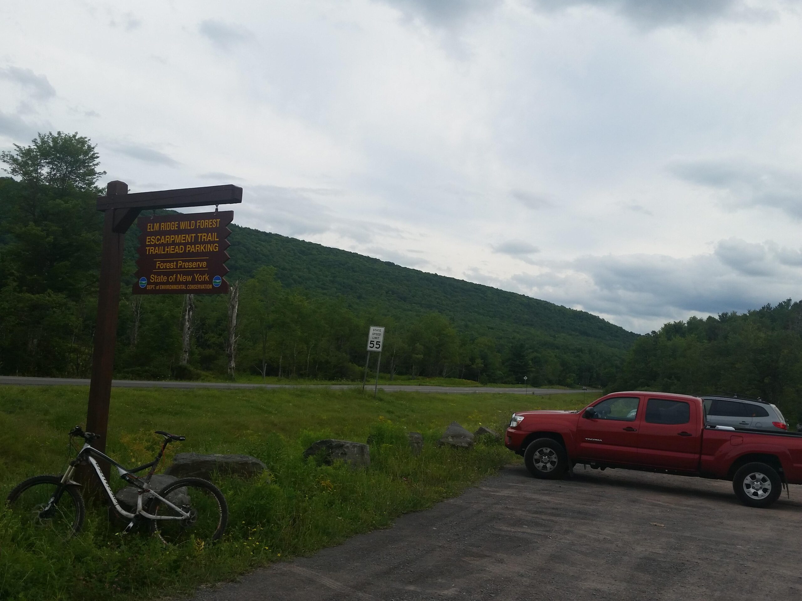 Specialized Stumpjumper FSR Comp EVO: A view of the Elm Ridge Wild Forest trailhead parking area, featuring a sign for the Escarpment Trail and a red pickup truck parked nearby. A mountain and cloudy sky are in the background, while a bicycle leans against the sign. The scene is set in a lush green landscape typical of a forested area in New York.