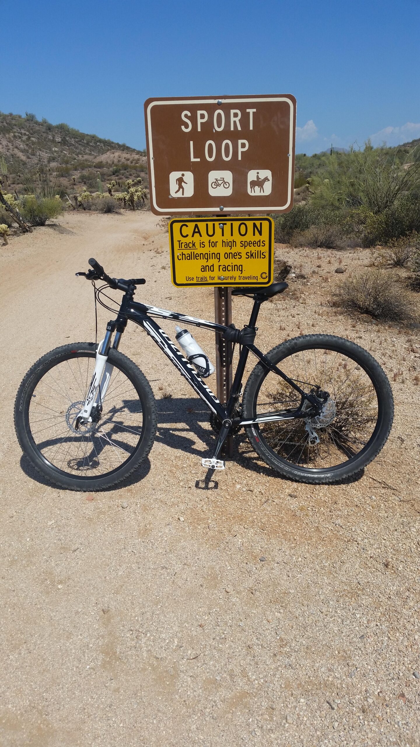 Specialized Pitch 650b: A mountain bike parked on a dirt trail next to a brown sign that reads "SPORT LOOP," indicating a loop trail for walking, biking, and horseback riding. A caution sign below warns that the track is for high speeds, challenging one's skills and racing. The background features a sunny sky and desert landscape with sparse vegetation.