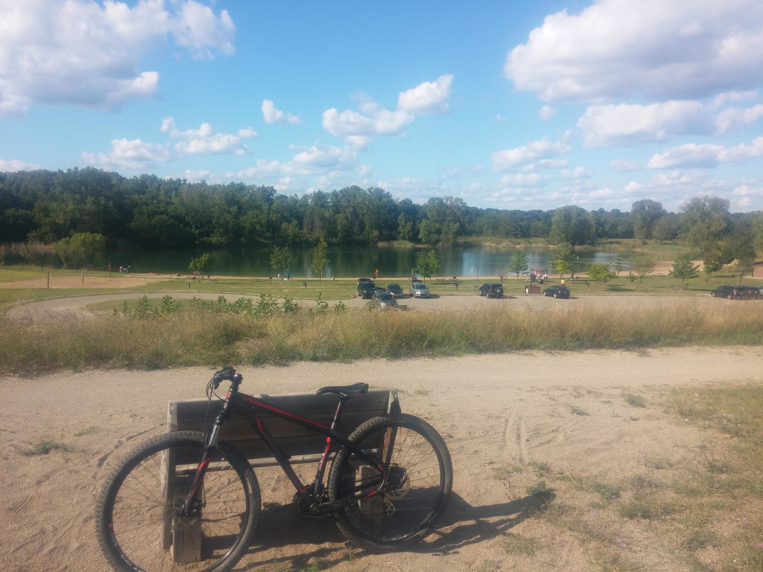 A mountain bike resting on a wooden bench in the foreground, with a sandy path leading to a serene lake in the background. The scene features lush greenery, scattered clouds in a blue sky, and a parking area filled with cars. People can be seen leisurely enjoying the area near the water. Island Lake mountain bike trail.