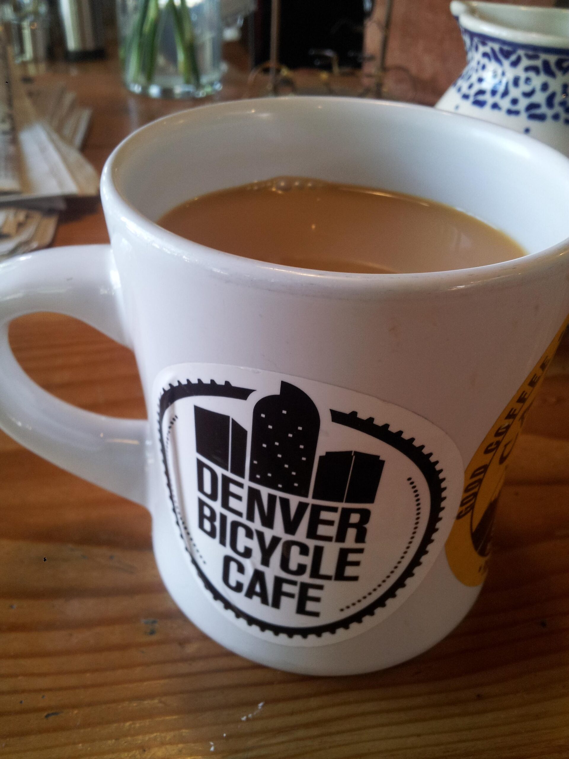 A white coffee mug with a black logo that reads "Denver Bicycle Cafe," filled with coffee. The mug is placed on a wooden table, with blurred background elements suggesting a café setting.