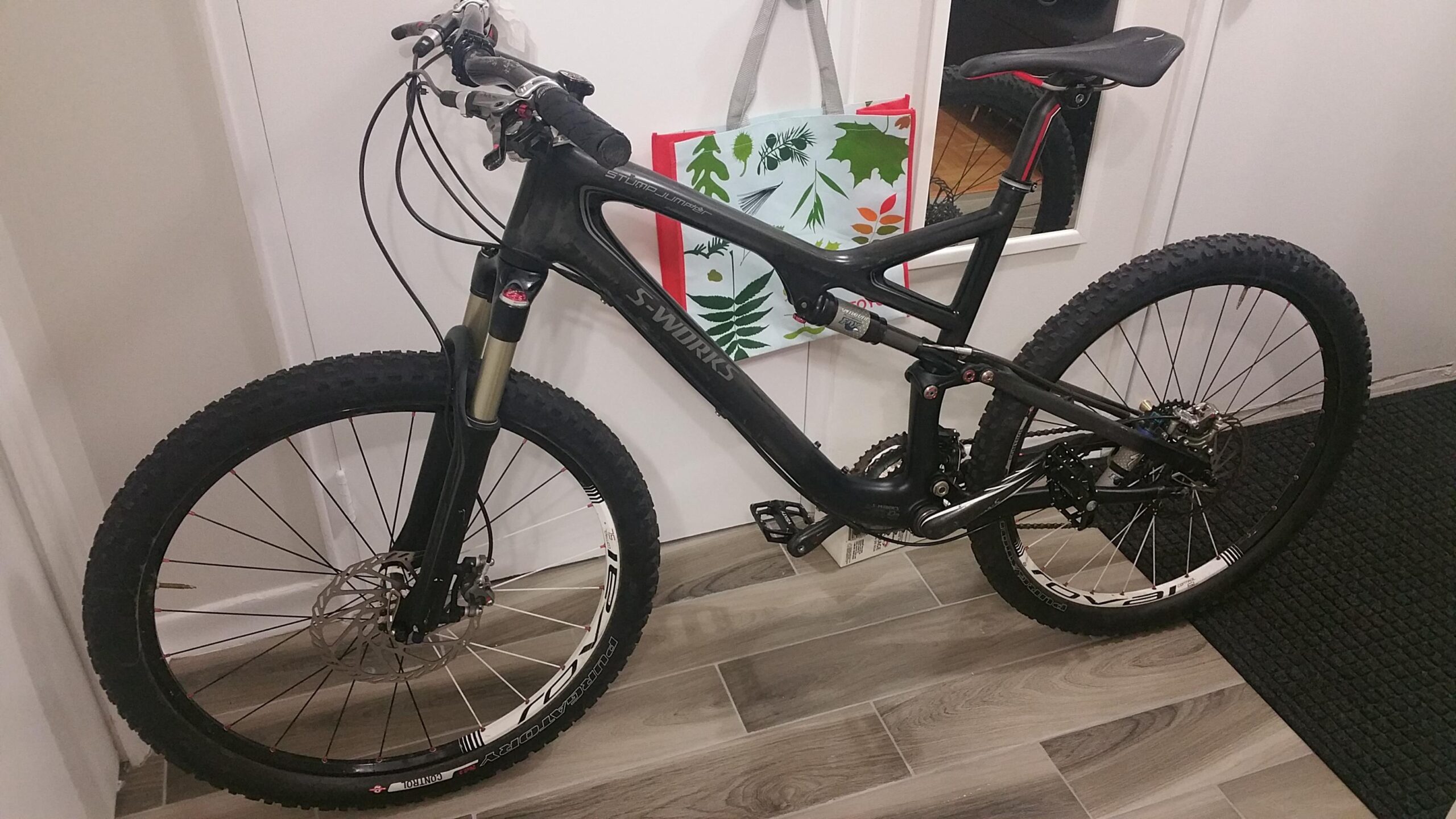 Specialized Stumpjumper: A black mountain bike with thick tires and a suspension system is parked next to a door. A colorful tote bag with leaf designs hangs on the wall behind it. The floor is made of light wood planks, and a dark textured mat is visible in the foreground.