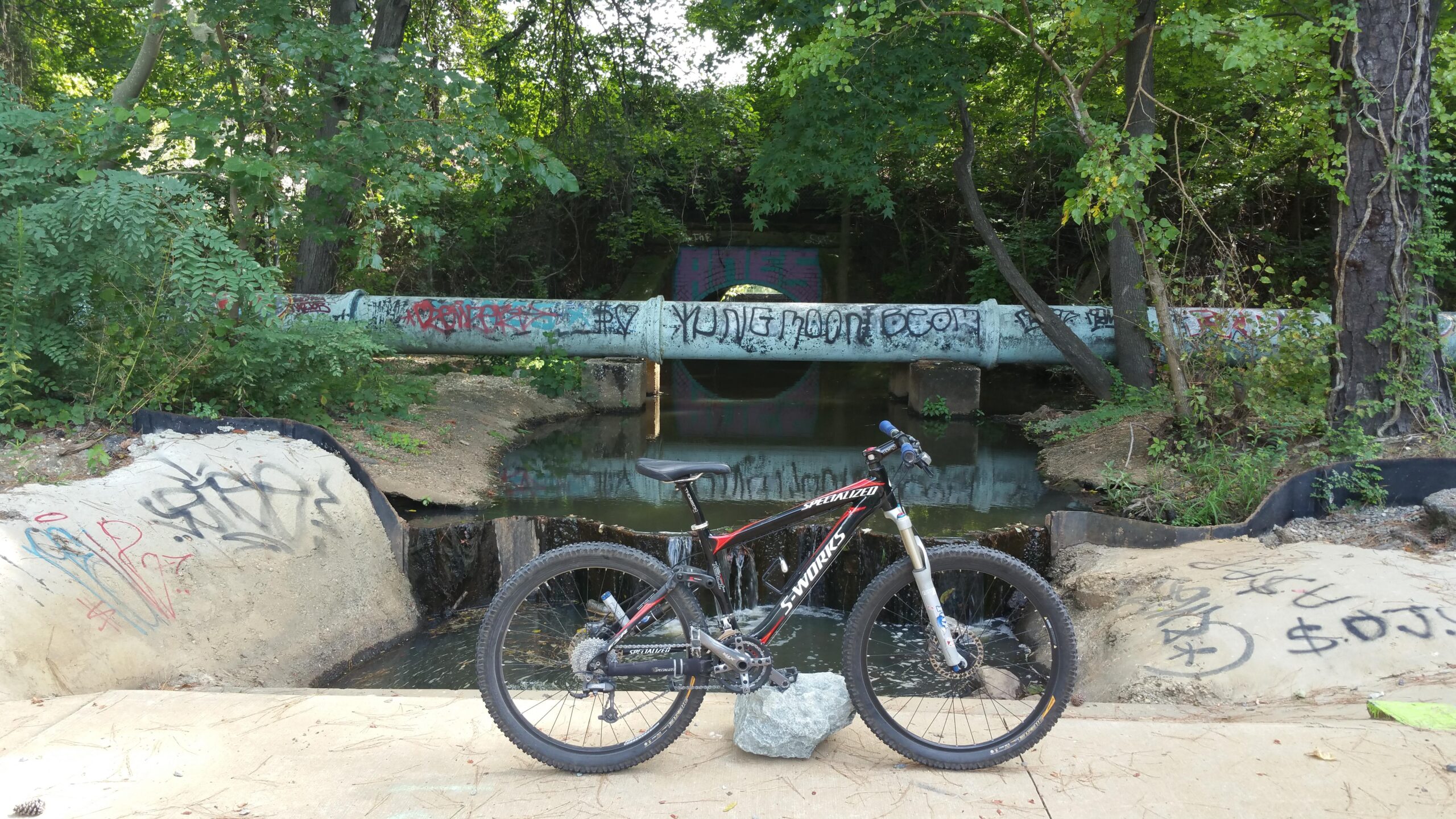 A mountain bike stands on a concrete path beside a small stream, with a graffiti-covered pipe in the background. The area is surrounded by dense green foliage, creating a natural yet urban atmosphere. The ground shows signs of graffiti art, adding to the vibrant and edgy feel of the scene. Lake Maury mountain bike trail.