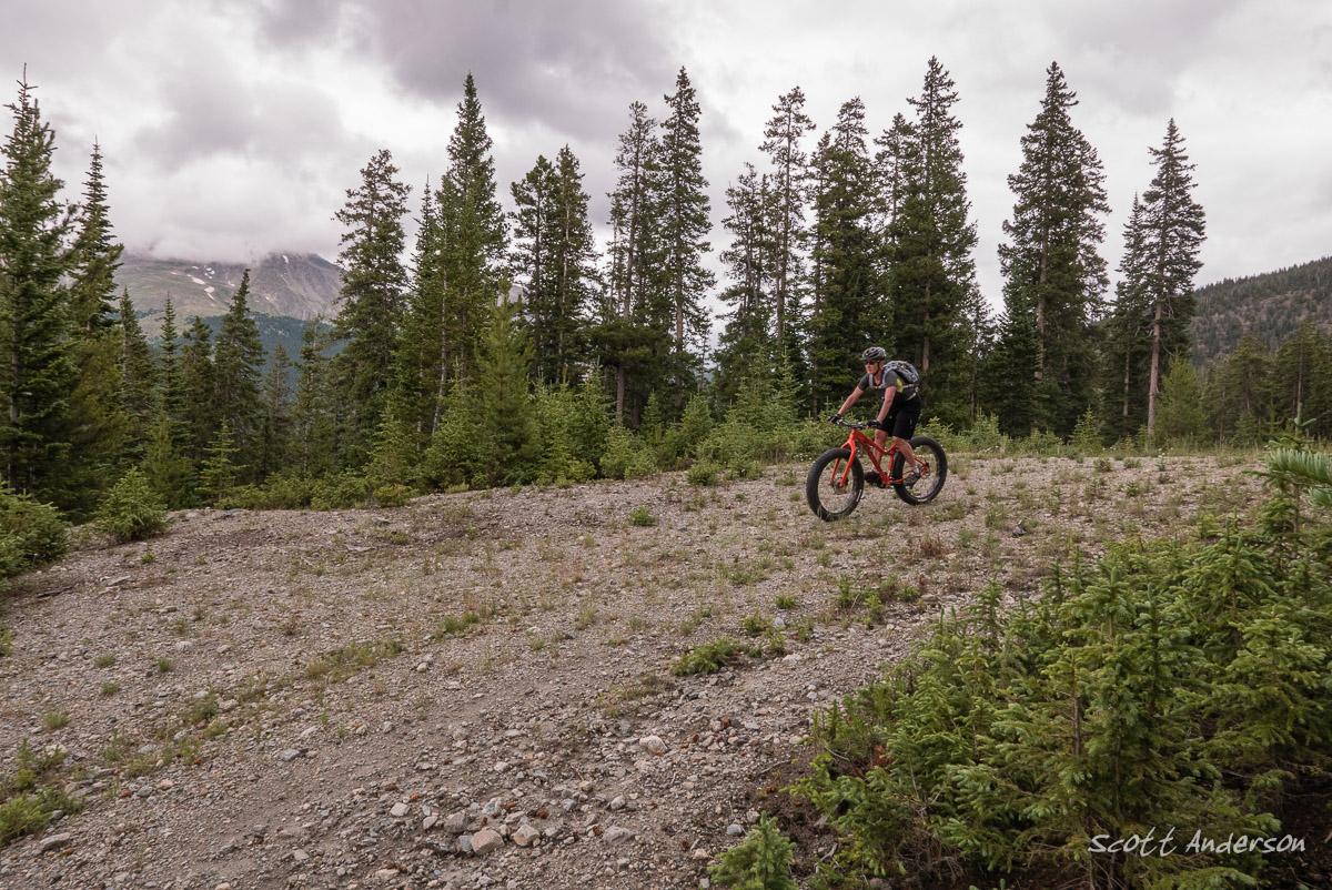 A mountain biker rides on a gravel path surrounded by tall evergreen trees, with a cloudy sky and distant mountains in the background. The biker is wearing a helmet and riding a bright red mountain bike. Cottonwood Pass Road mountain bike trail.