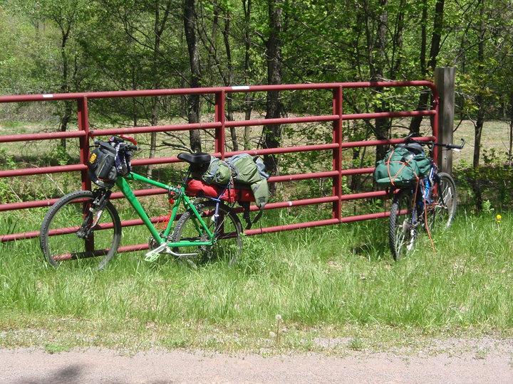 Diamondback Response: Two bicycles are parked against a red metal gate, surrounded by lush green grass and trees. The bicycle on the left is green and has bags attached to the front and rear, while the bicycle on the right is blue and also carries additional bags. The scene is set in a natural outdoor environment, indicating a moment of rest during a biking trip.