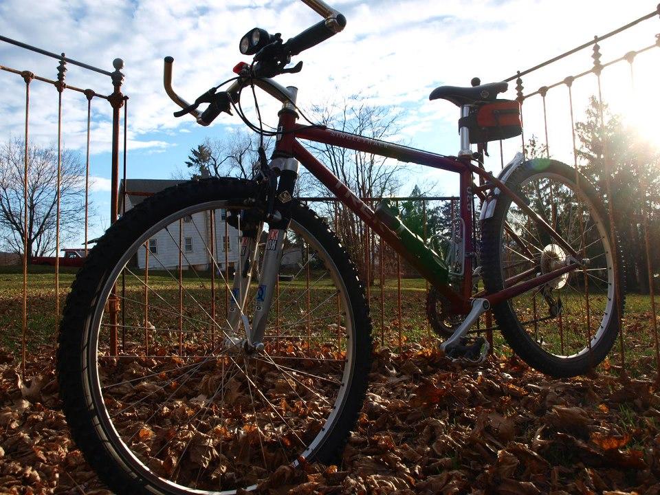Trek 930: A mountain bike resting on a bed of fallen leaves, with a rustic iron fence in the background. The sun is setting, casting a warm light, and a simple house can be seen in the distance.