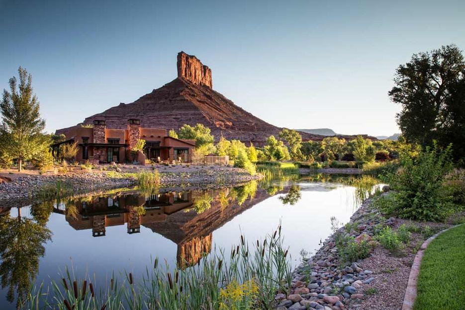 Scenic view of a tranquil river reflecting a towering rock formation, surrounded by lush greenery and a rustic building. The landscape showcases the beauty of nature with a clear sky above.