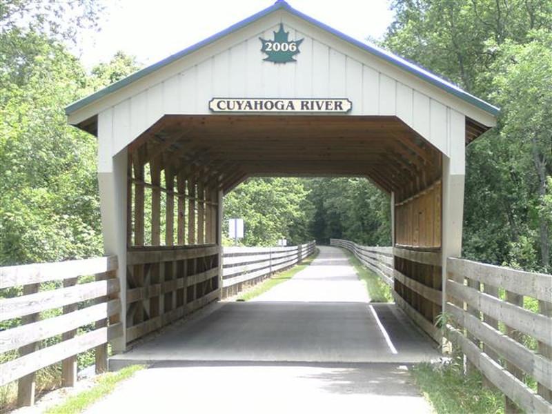 Covered bridge with "Cuyahoga River" sign at the entrance, surrounded by greenery, leading to a pathway. The bridge features wooden beams and sides, with a clear view of the trail extending beyond. A sign indicating the year 2006 is displayed on the roof. Maple Highlands Trail-Mountain Run Station mountain bike trail.