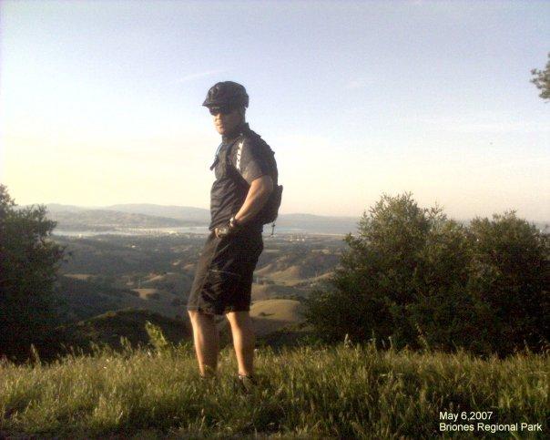 A person in athletic attire stands on a grassy hillside overlooking a scenic landscape at Briones Regional Park. The individual wears a helmet and appears to be enjoying the view, surrounded by trees and hills under a clear sky. The date captures the moment as May 6, 2007. Briones Regional Park mountain bike trail.