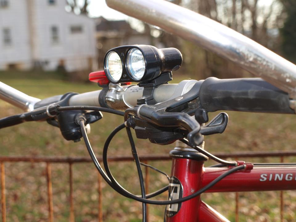 Trek 930: Close-up view of a bicycle handlebar featuring a dual LED headlight, brake lever, and gear shifter. The background is an outdoor setting with blurred greenery and an out-of-focus white building.