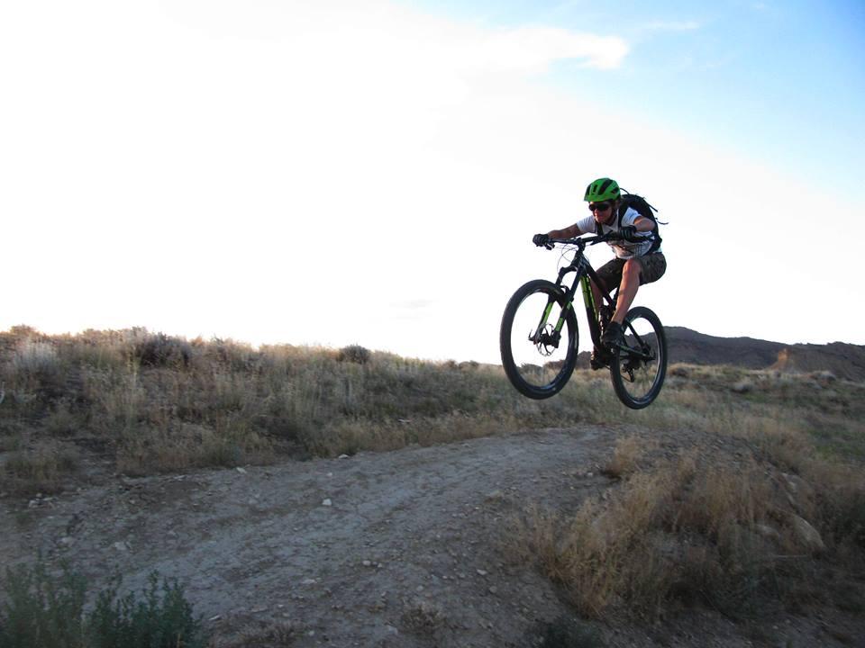 A mountain biker wearing a green helmet and a backpack is airborne above a dirt path, performing a jump on a hillside during daylight. The background features a clear sky and rolling hills with sparse vegetation.